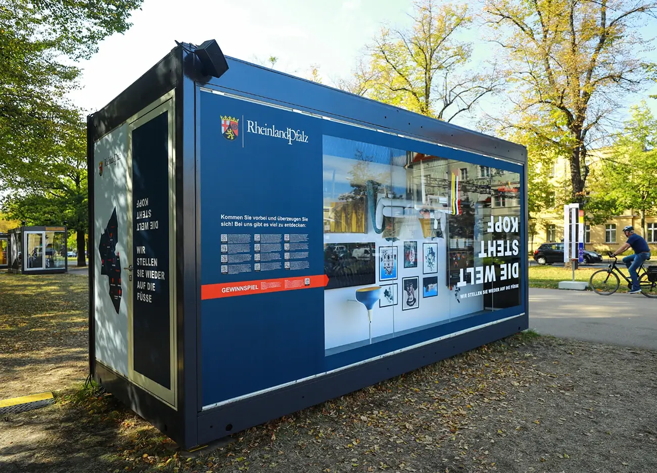 Frontansicht des Pavillon für Rheinland-Pfalz anlässlich des Tag der Deutschen Einheit, in dem ein auf dem Kopfstehendes Wohnzimmer aufgebaut ist, mit der Aufschrift "Die Welt steht Kopf - Wir stellen sie wieder auf die Füsse"